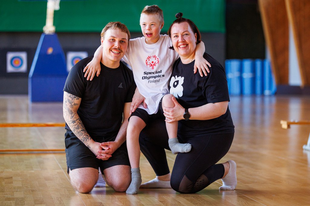 Iloinen perheleiriläinen, poikaoletettu, yhteiskuvassa kahden perheenjäsenensä kanssa. Päällään hänellä valkoinen SO t-paita, jossa punainen SO-logo.